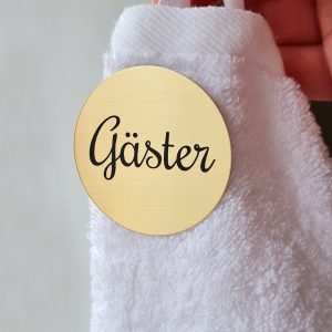 Vit handduk och mässingsskylt med graverad text. Gäster.
