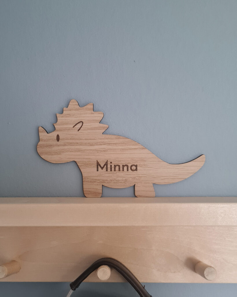 Skylt till barnrum med formen av en dinosourie. Graverat namn som visar var kläder och väskor ska hängas.